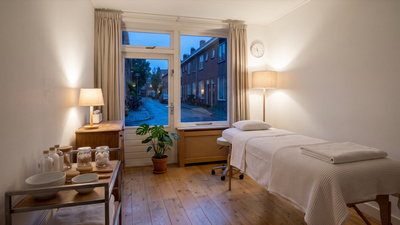 De lege behandelkamer van een salon aan huis in de avond, met warm lamplicht en de straat buiten