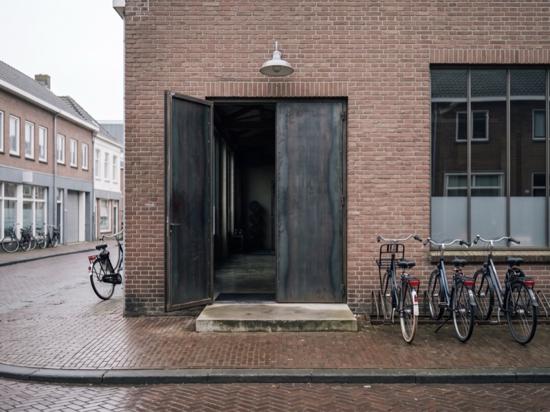 De stalen deur van de loods staat open aan een bakstenen straat, met fietsen ernaast